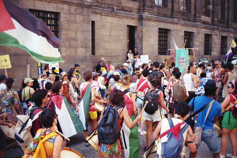 The image portrays a lively street scene where a large crowd of people has gathered. They are holding flags and signs, suggesting they might be protes