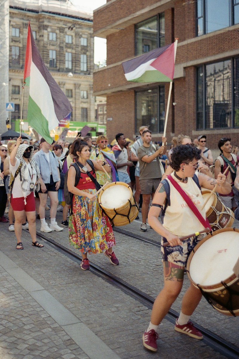 This image captures a vibrant street scene. There is a group of people marching down the road with their drums. They are waving flags, indicating they