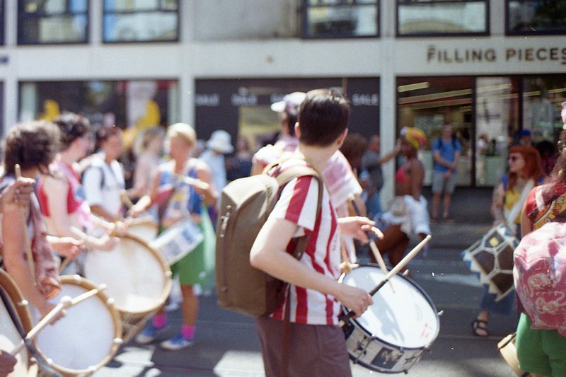 A group of people are standing together in the street. They seem to be part of a marching band or festival, as suggested by their colorful attire and
