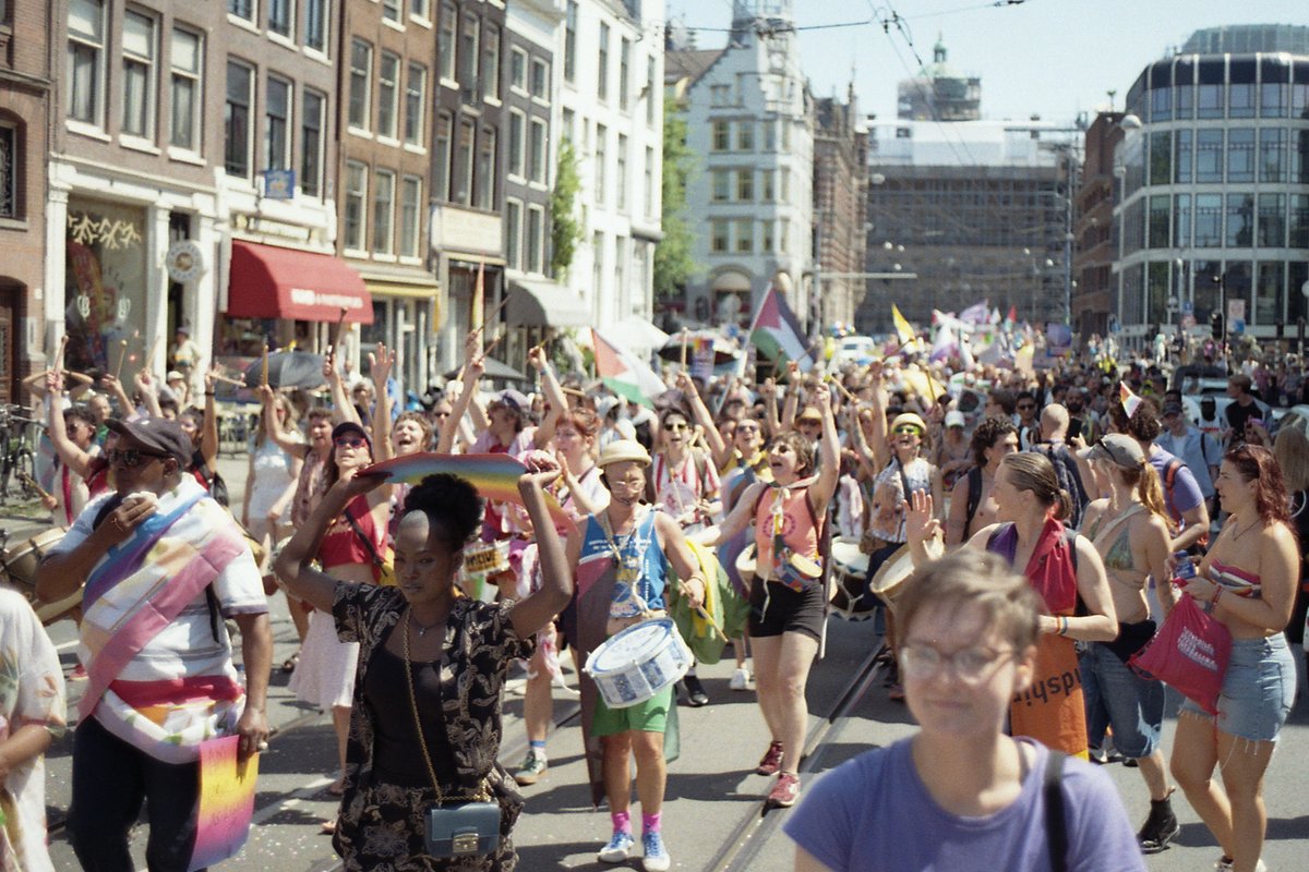 A vibrant city street filled with people of various ages and outfits. They are gathered in celebration, waving flags and banners. Some of the individu