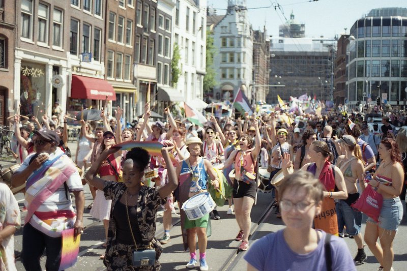A vibrant city street filled with people of various ages and outfits. They are gathered in celebration, waving flags and banners. Some of the individu