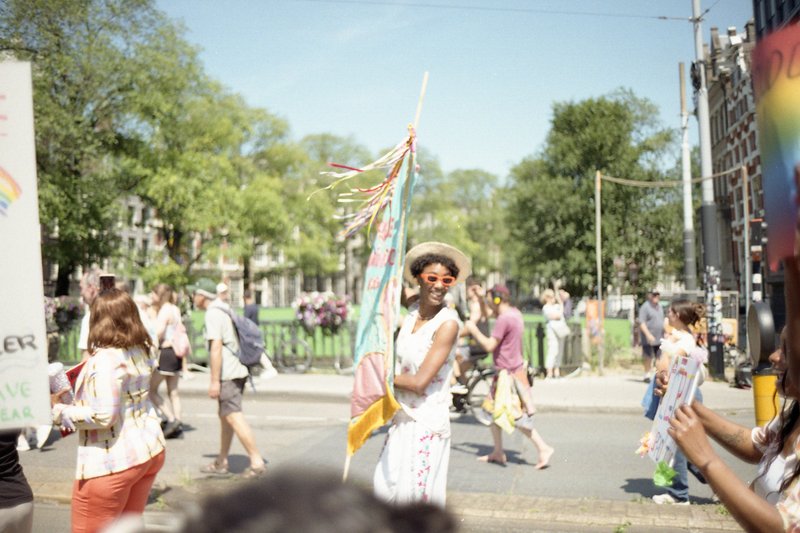 The image captures a lively scene of a parade. At the center, a person stands out with their colorful attire, possibly representing LGBTQ+ pride or a