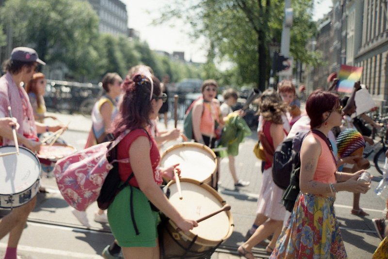 The image shows a lively scene of a parade or march. A group of people is seen walking down a city street. Some individuals carry backpacks and musica
