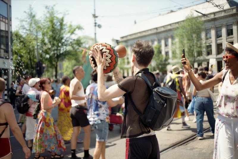 The photo depicts a lively street scene with many people gathered. A prominent person is seen wearing a mask and holding a drum, possibly participatin
