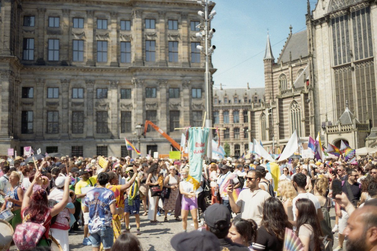 The image depicts a lively street festival. A large crowd of people are gathered, enjoying the festivities with flags flying in the background and col