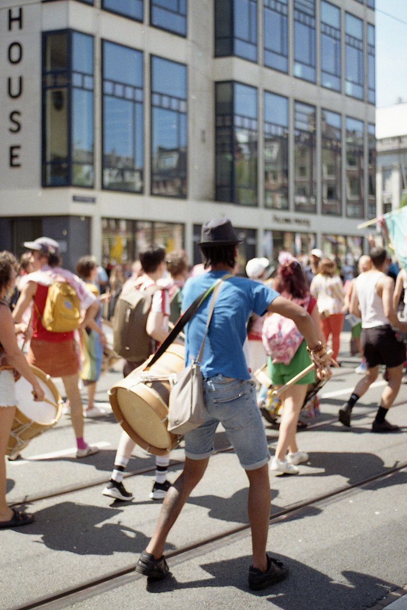 A lively street festival. People are marching down the street, with some carrying drums and banners. One man is prominently visible, dressed casually