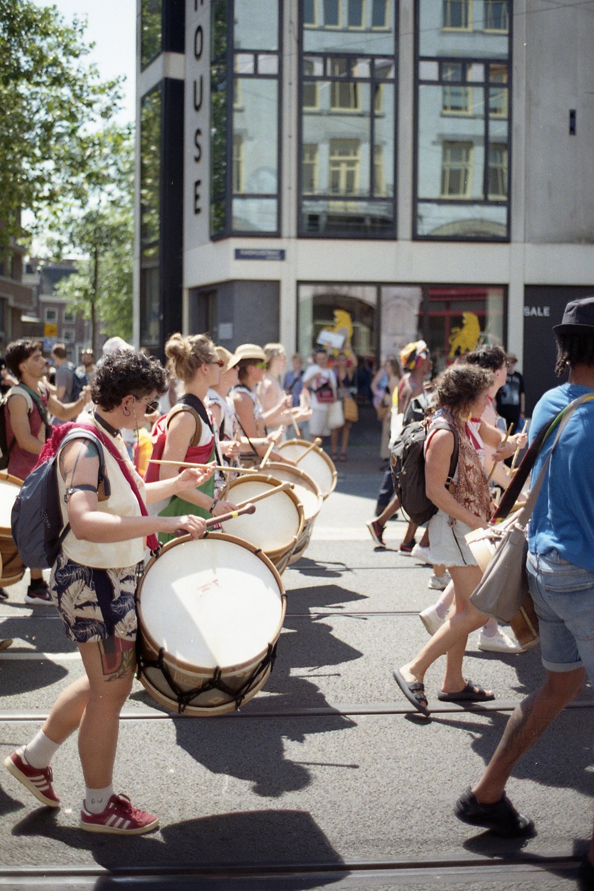 A lively street scene featuring a marching band, with drummers at the forefront. The band members are carrying large percussion instruments and some e