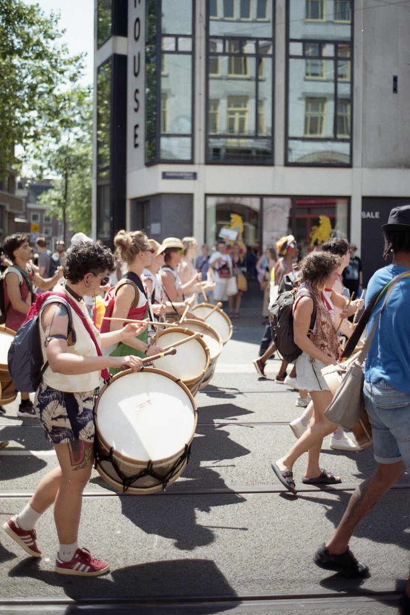 A lively street scene featuring a marching band, with drummers at the forefront. The band members are carrying large percussion instruments and some e
