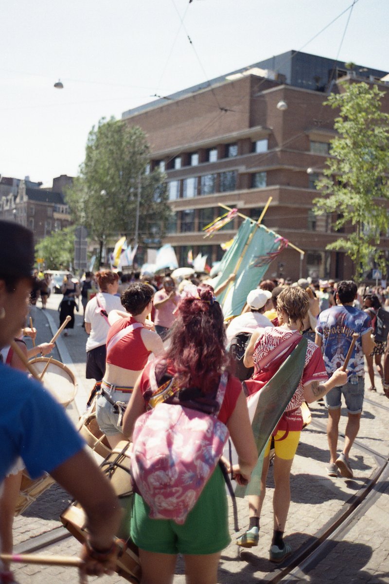A group of people marching down a street, holding banners and flags. The atmosphere appears to be festive or celebratory. They're walking on a sidewal