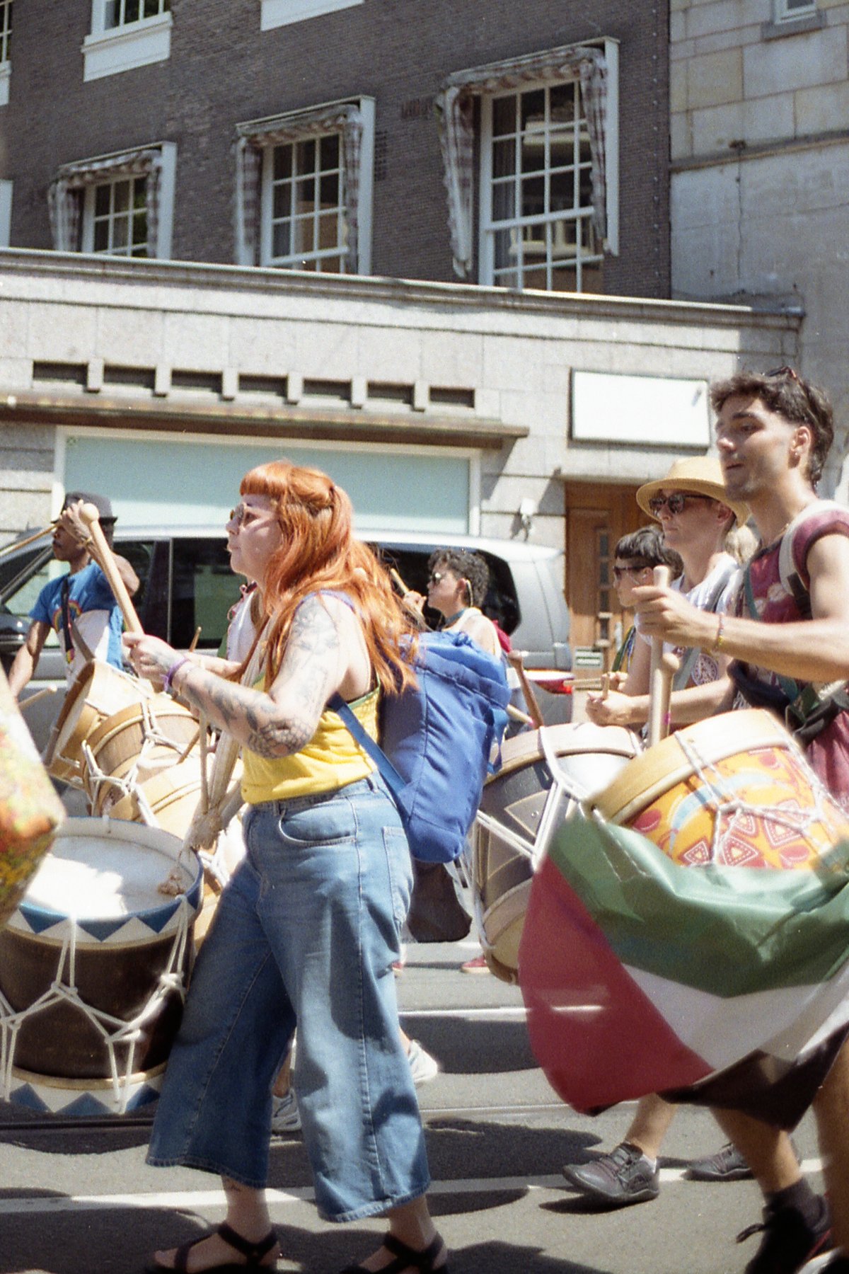 A vibrant street scene with several people playing musical instruments and marching. The crowd is diverse, including individuals holding various percu