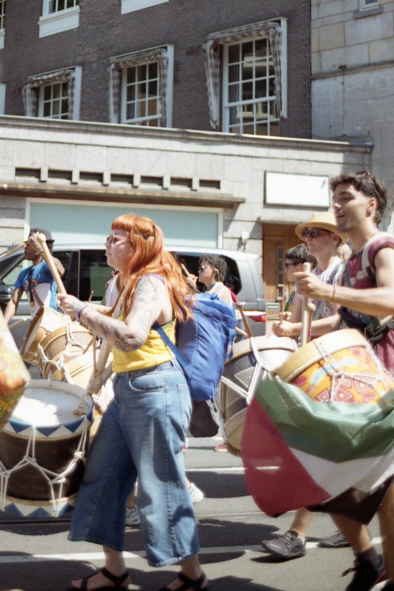 A vibrant street scene with several people playing musical instruments and marching. The crowd is diverse, including individuals holding various percu