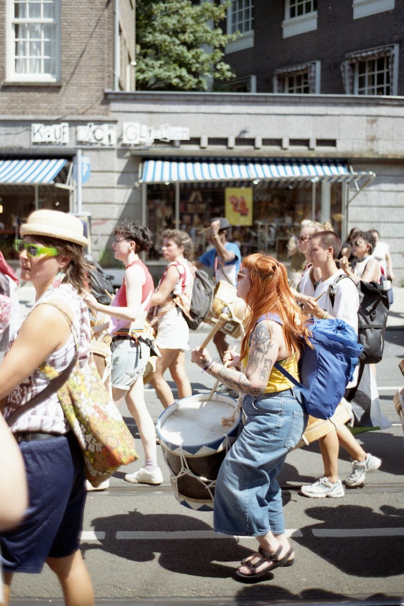 A lively street scene featuring a parade with people walking around and playing musical instruments. The image depicts a variety of individuals partic