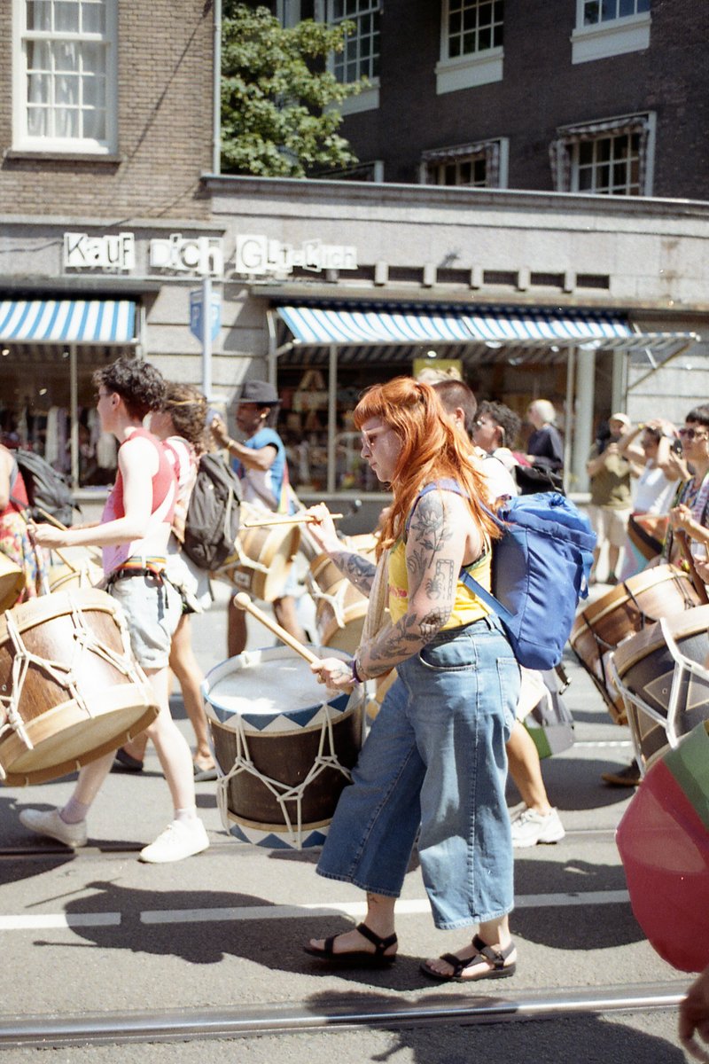 The image is a photograph taken from a distance during the day. It features a large group of people marching down a street, some carrying drums and ot