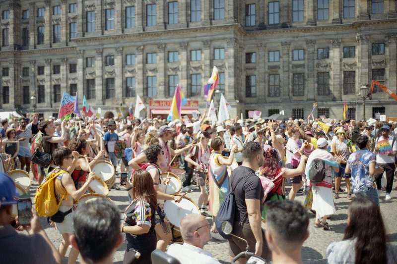 The image depicts a vibrant street scene. A large crowd has gathered, with many people holding signs and banners. There are also several musicians pla