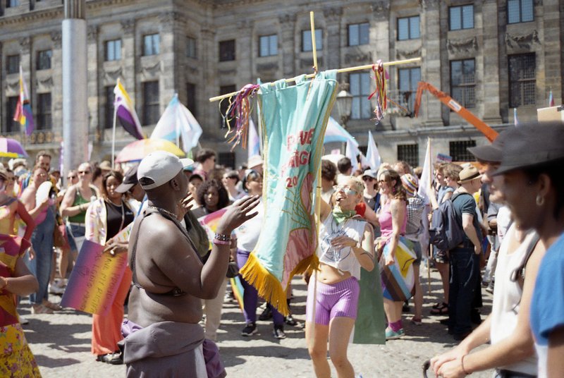 A vibrant gathering of people on a street corner. Some participants are holding flags and banners. The atmosphere seems festive with individuals dress