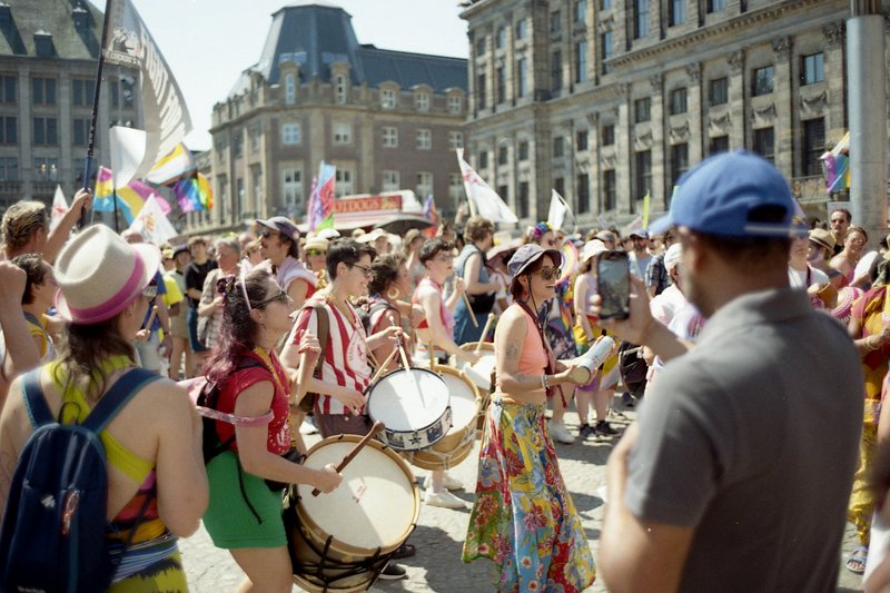 A lively scene of a parade on a street. People are marching, some holding instruments like drums and tambourines. The crowd includes a diverse group o