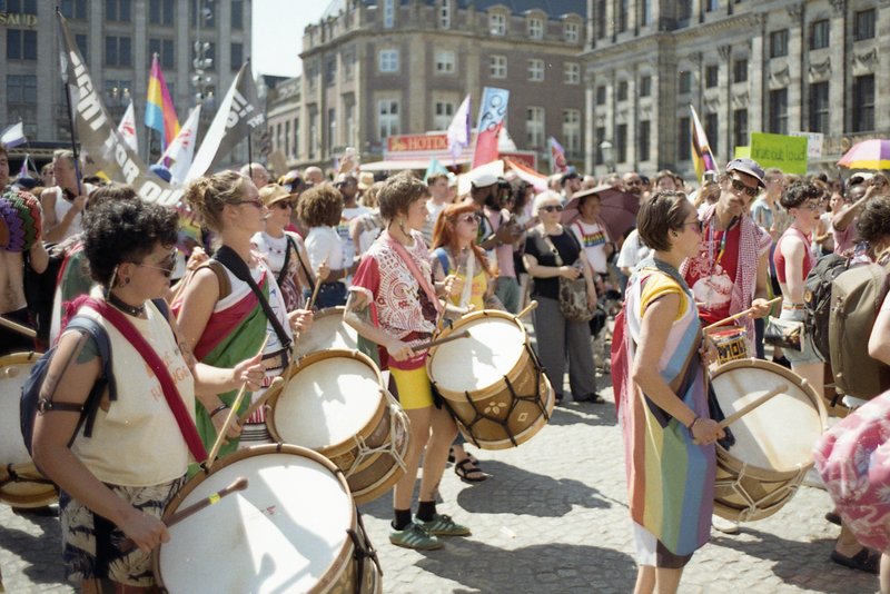 The image depicts a large gathering of people participating in what appears to be a protest or parade. In the foreground, individuals are holding drum