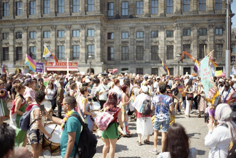 This image captures a vibrant scene at a public square. A large crowd of people is gathered, with many carrying colorful flags and banners. Some indiv