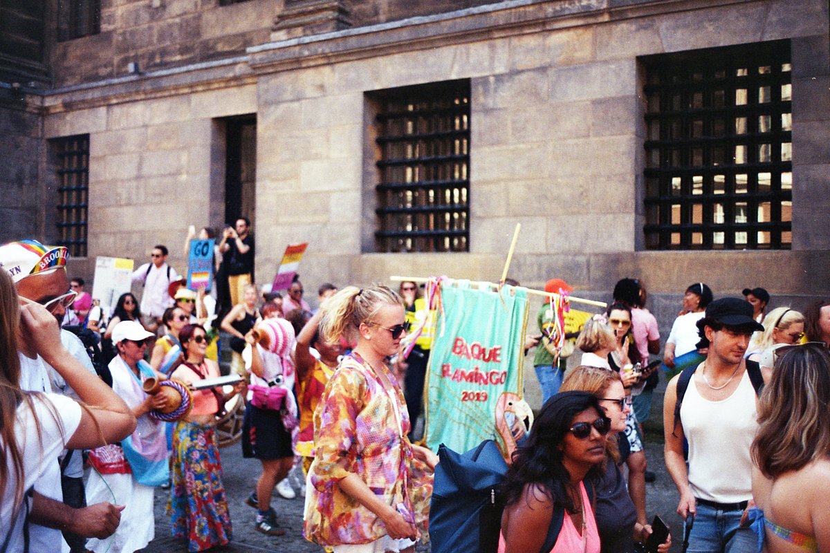 A lively street scene with a crowd of people, many holding signs and banners. The group seems to be celebrating or protesting outside a large building