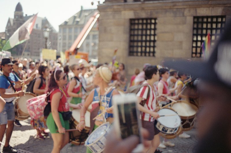 A group of people playing music outdoors. There are drummers and other musicians wearing colorful attire. The scene suggests a public event or celebra