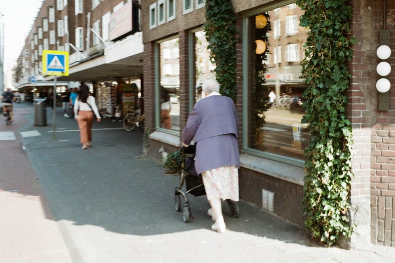 A woman with a walker walks down the street. She's dressed warmly and is carrying a handbag. The setting appears to be a city street with shops lining