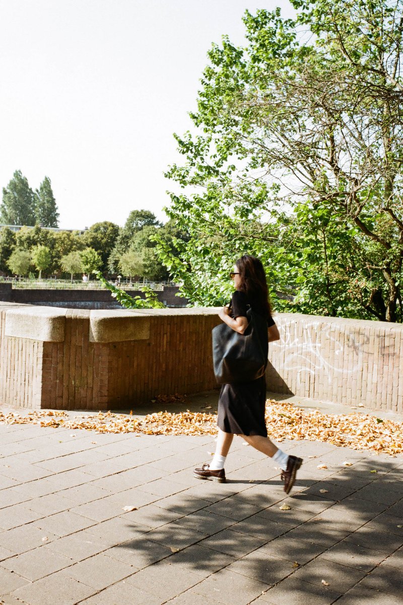 Person walking alone, near trees. Brick sidewalk. This is an urban setting. The person is carrying a backpack and dressed casually, appropriate for ou