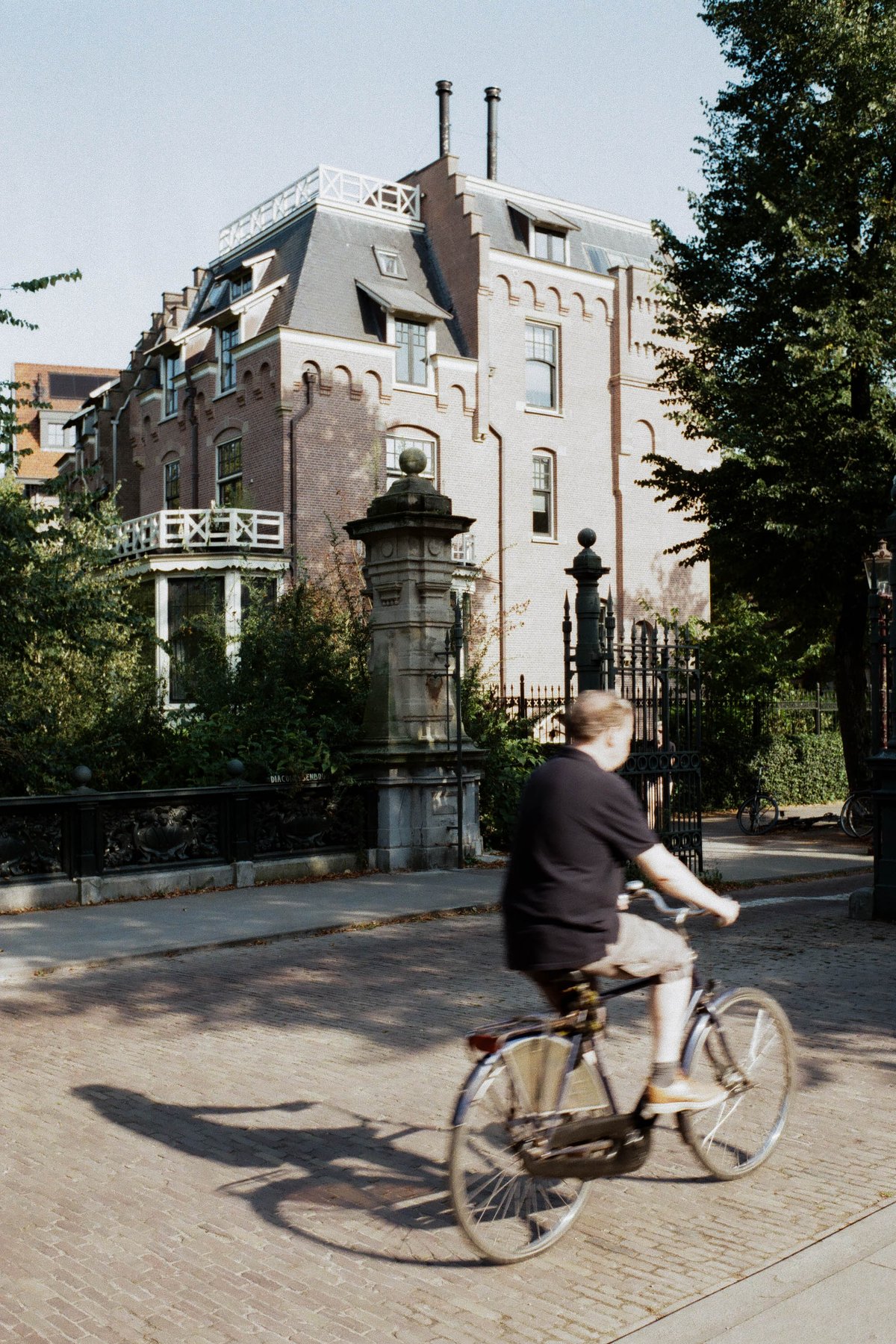 The image shows a person riding a bicycle on a paved street. The cyclist appears to be in motion. To the left of the cyclist is a two-story building w