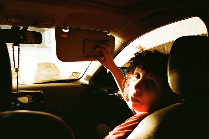 A black and white photo of a person sitting in the back seat of a car. The individual is holding onto the backrest, looking out through the open windo