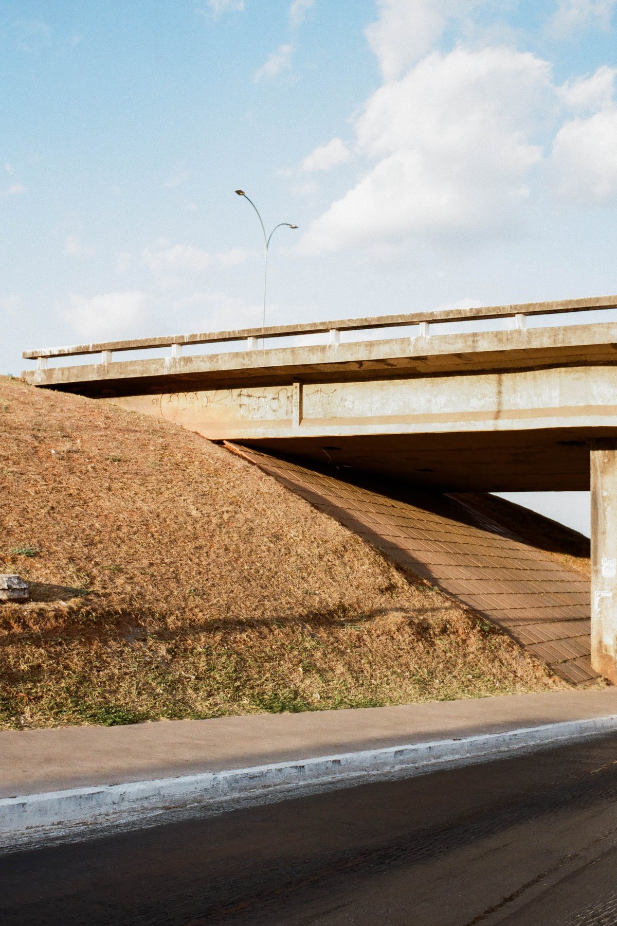 A color photograph of a single-lane concrete overpass during daytime. The overpass is brown and stretches across the image. It's set against a backdro