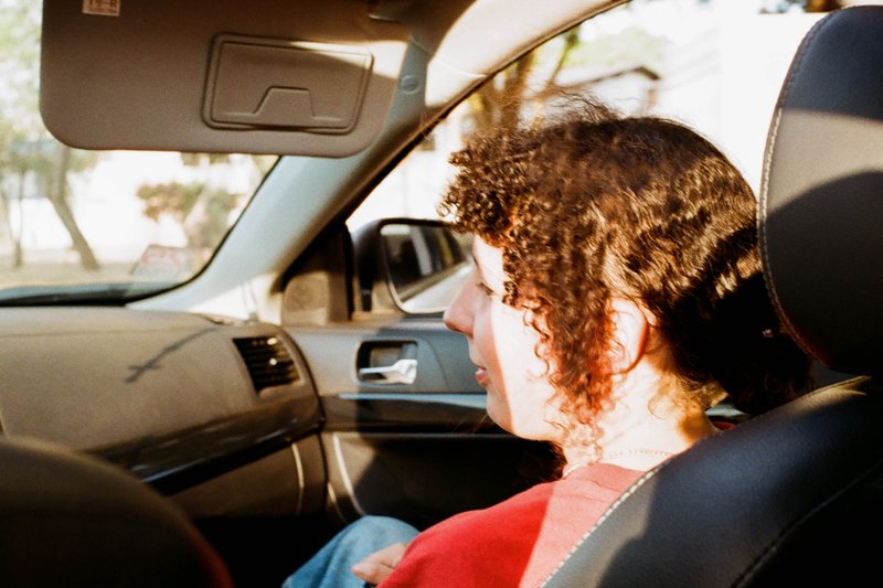 A woman sits in the passenger seat of a car. She has curly hair and is wearing glasses. The interior of the vehicle features a black leather upholster