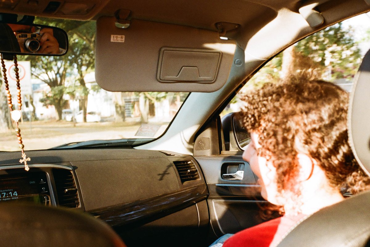 A man is seated inside a car, with the window open. The interior of the car is visible, and it appears to be parked during the daytime. Outside, there