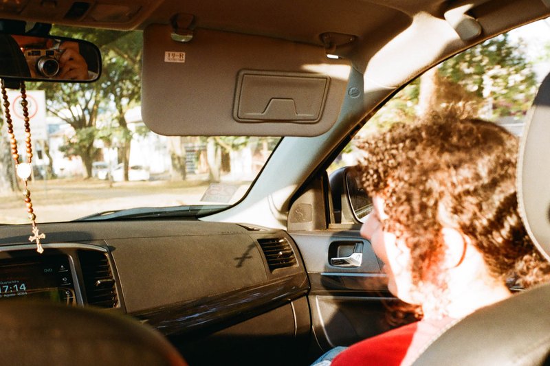 A man is seated inside a car, with the window open. The interior of the car is visible, and it appears to be parked during the daytime. Outside, there
