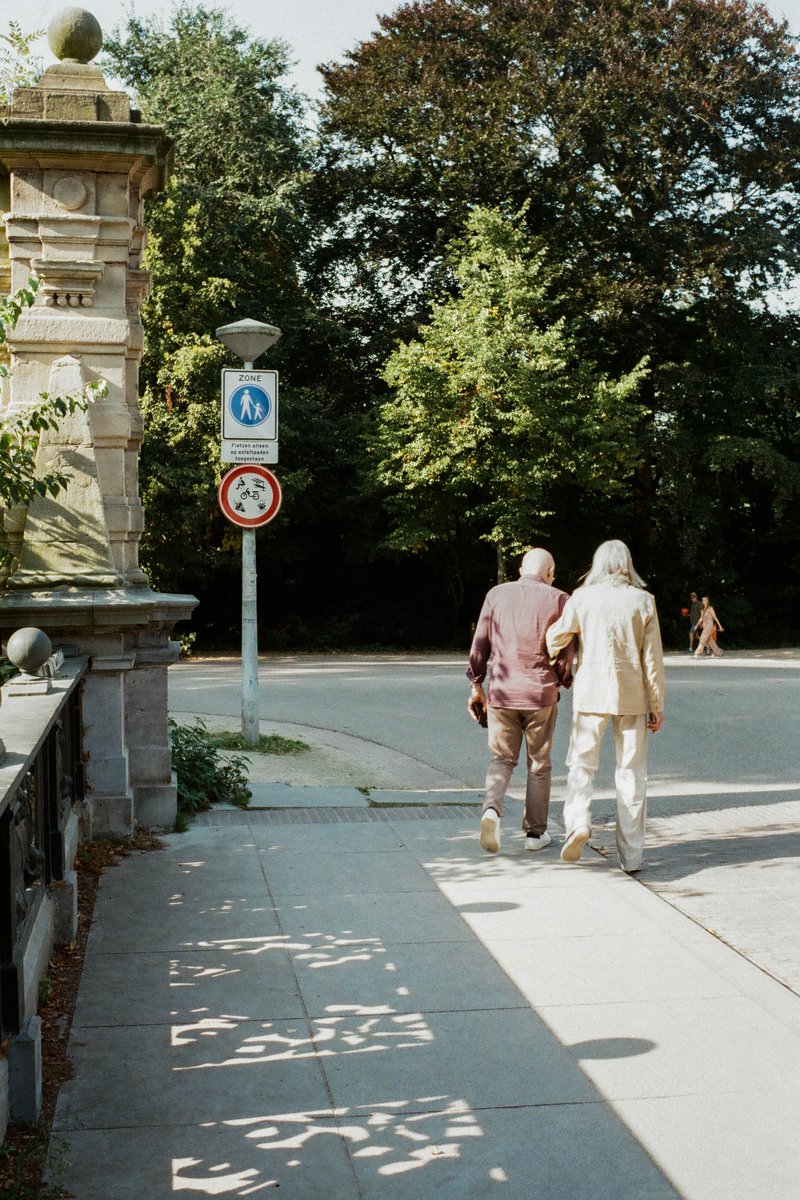 Two elderly people walking side by side down a sidewalk. A man and woman with white hair. Both are dressed casually and are wearing glasses. The woman