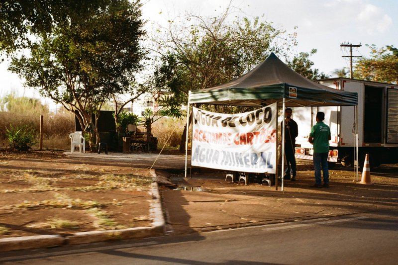 The image depicts a bustling scene on a street where a food stall is set up under a tent. Various people are standing around the stall, engaging in co