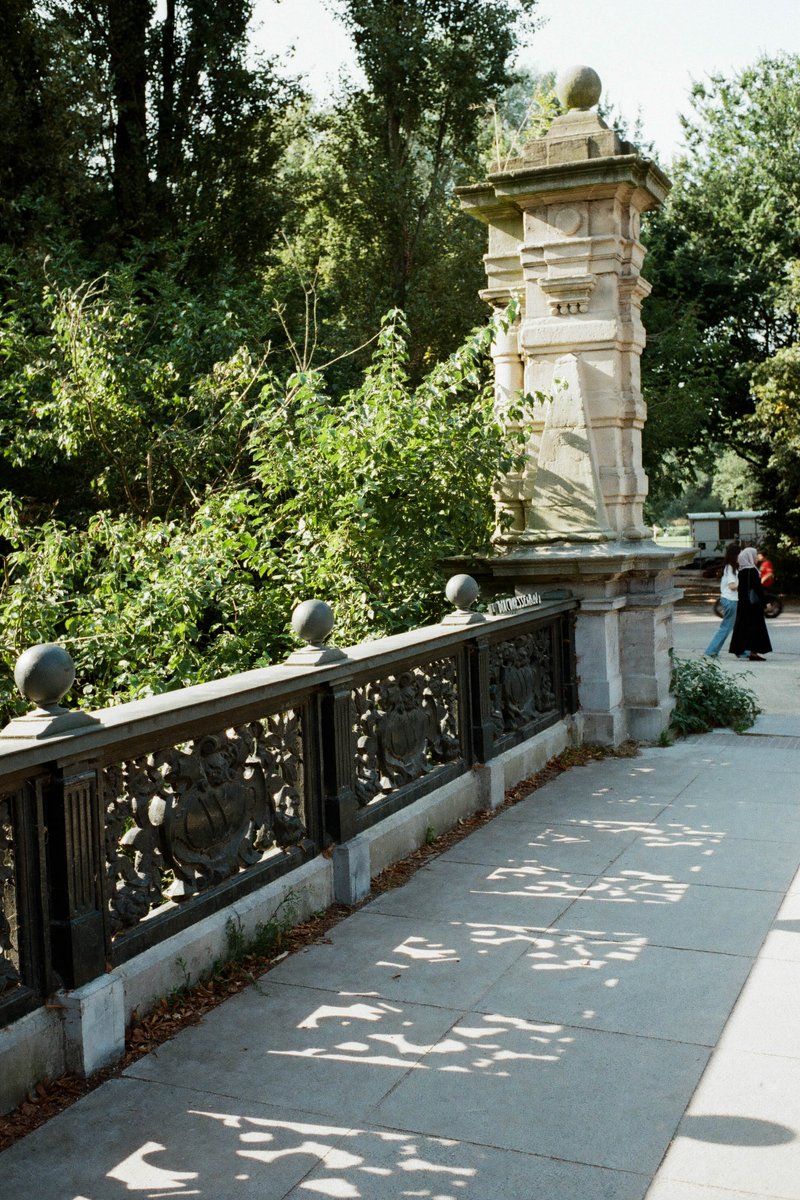 The image depicts a serene scene at the park. A stone fence runs along the side of a pathway, providing a sense of boundary and safety. The pathway, l