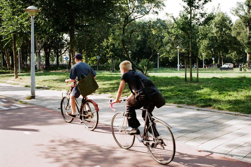 A vibrant scene of two cyclists riding down a city street lined with trees. Both are equipped with backpacks and one has a handbag. The backdrop featu