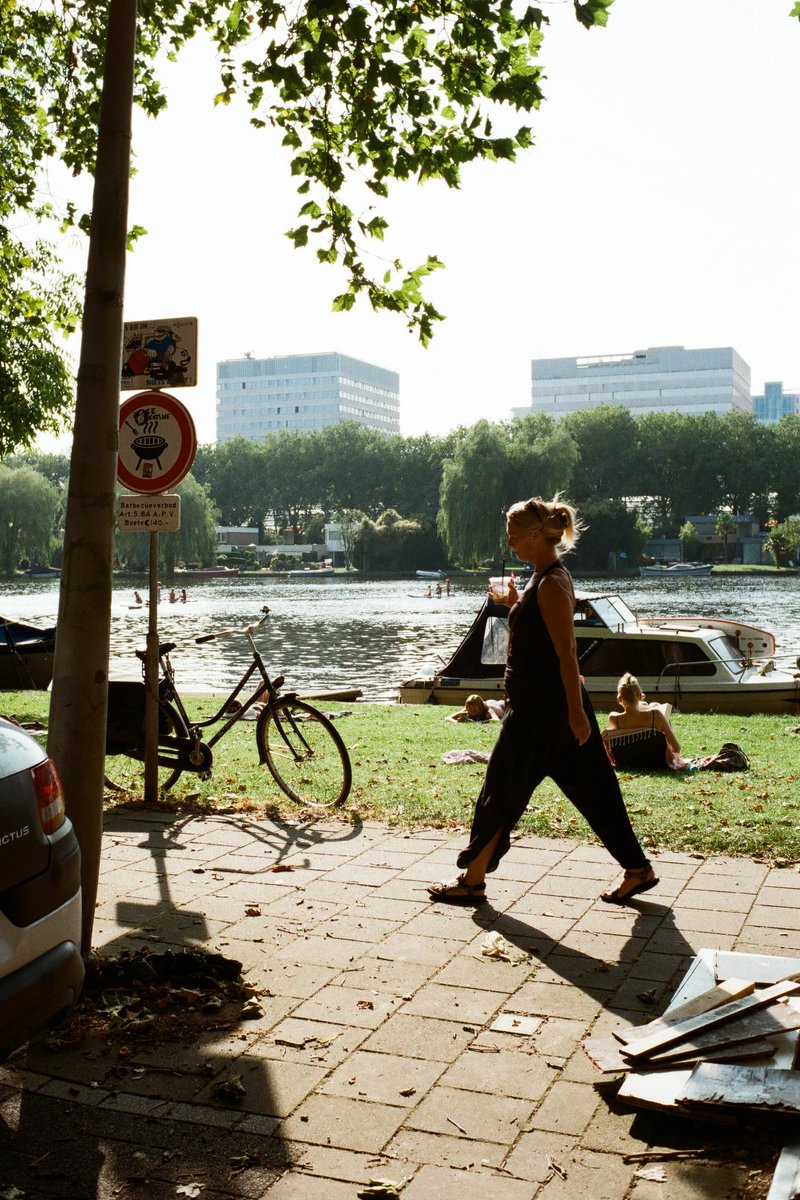 The image depicts a lively scene from a city park. A woman is strolling along the path, with a beautiful river flowing alongside. She seems to be enjo