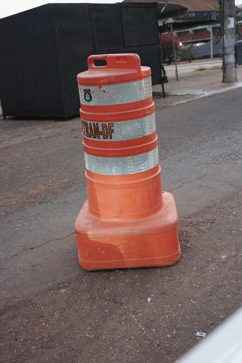 The image shows an orange construction barrel parked on the side of a road. There's a visible "Caution" sign indicating work zone. In the background,