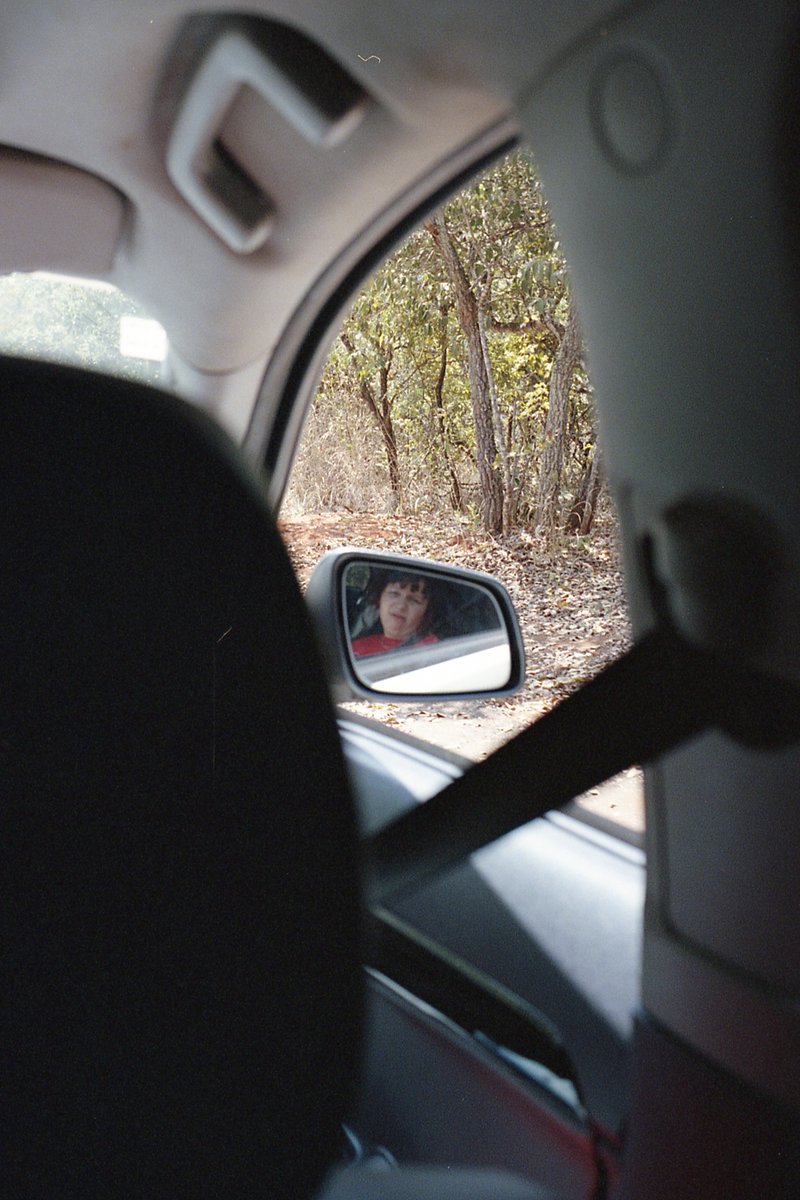 This is a black and white photo showing the interior of a car. Through the window on the left side of the vehicle, you can see a person sitting in the