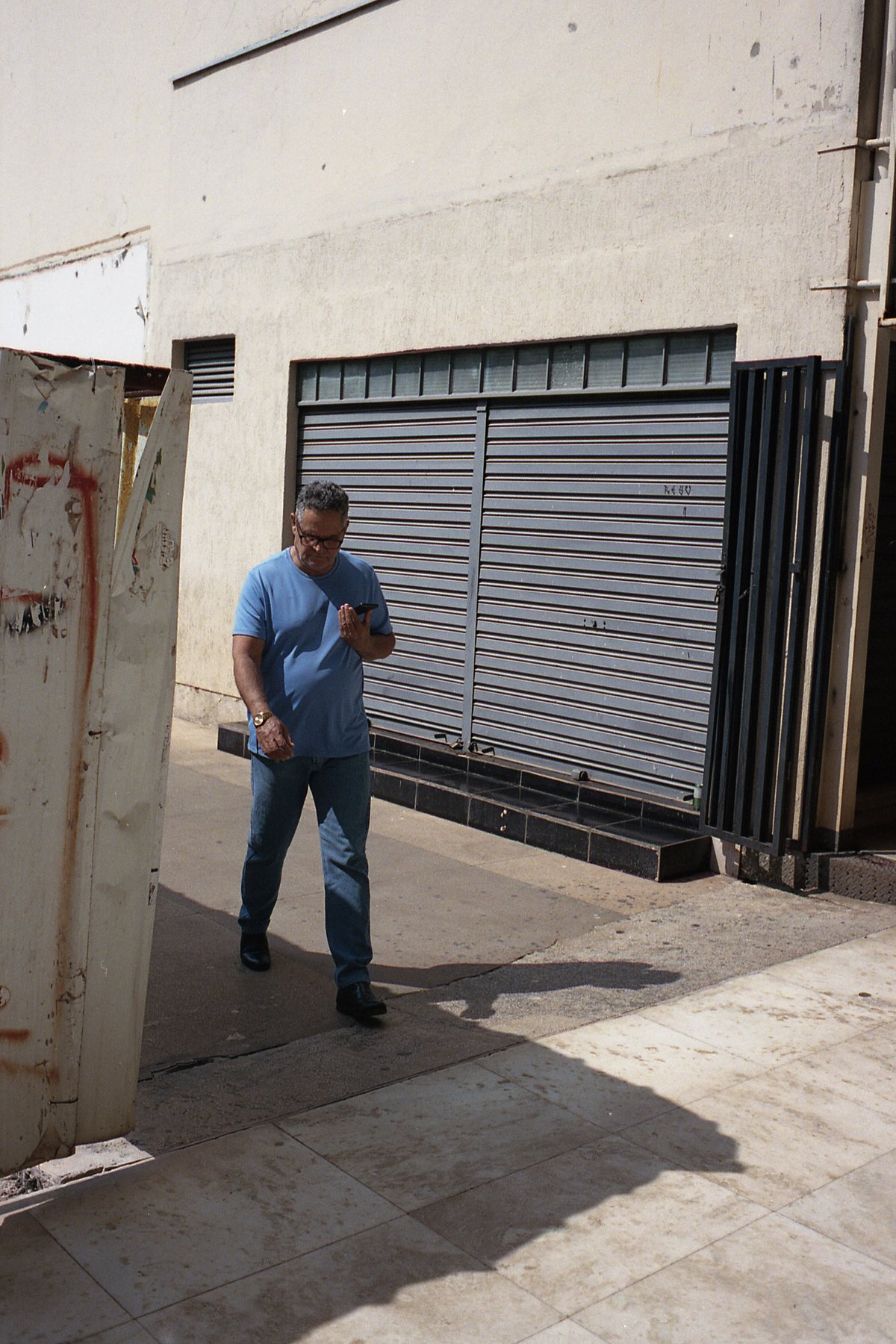A man stands in front of a closed building, looking at his phone. The building has a shuttered window and a sign on the side.