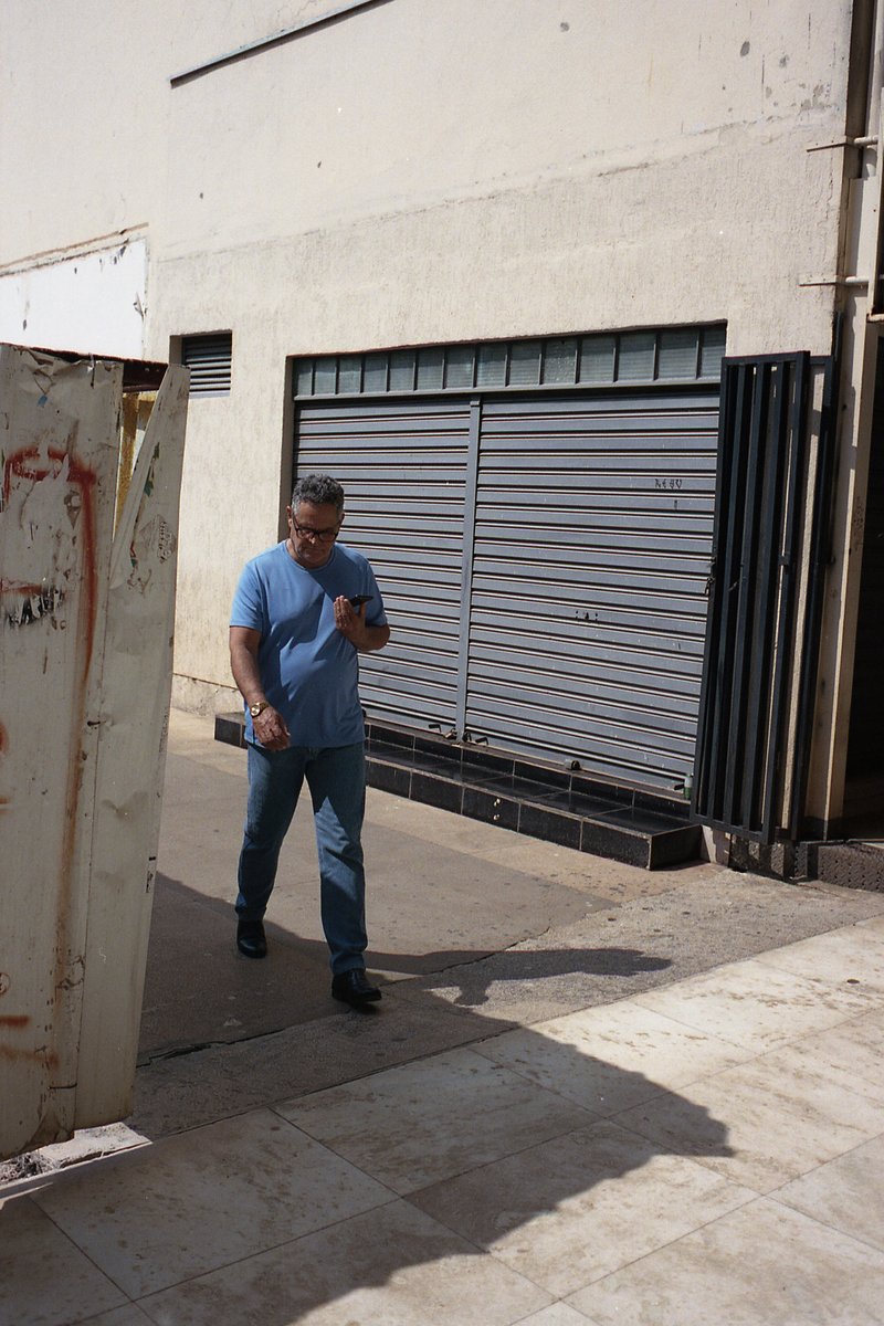 A man stands in front of a closed building, looking at his phone. The building has a shuttered window and a sign on the side.