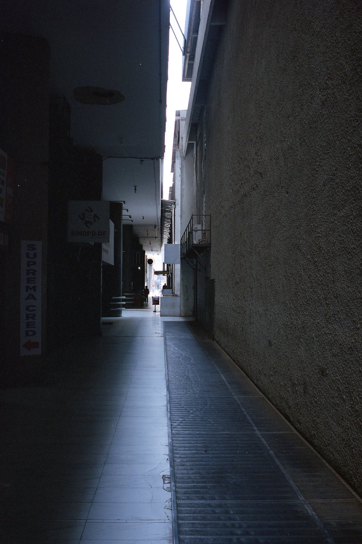 This is a black and white photo of an indoor corridor. The corridor appears to be made of cement or concrete and is lined on both sides by buildings. 