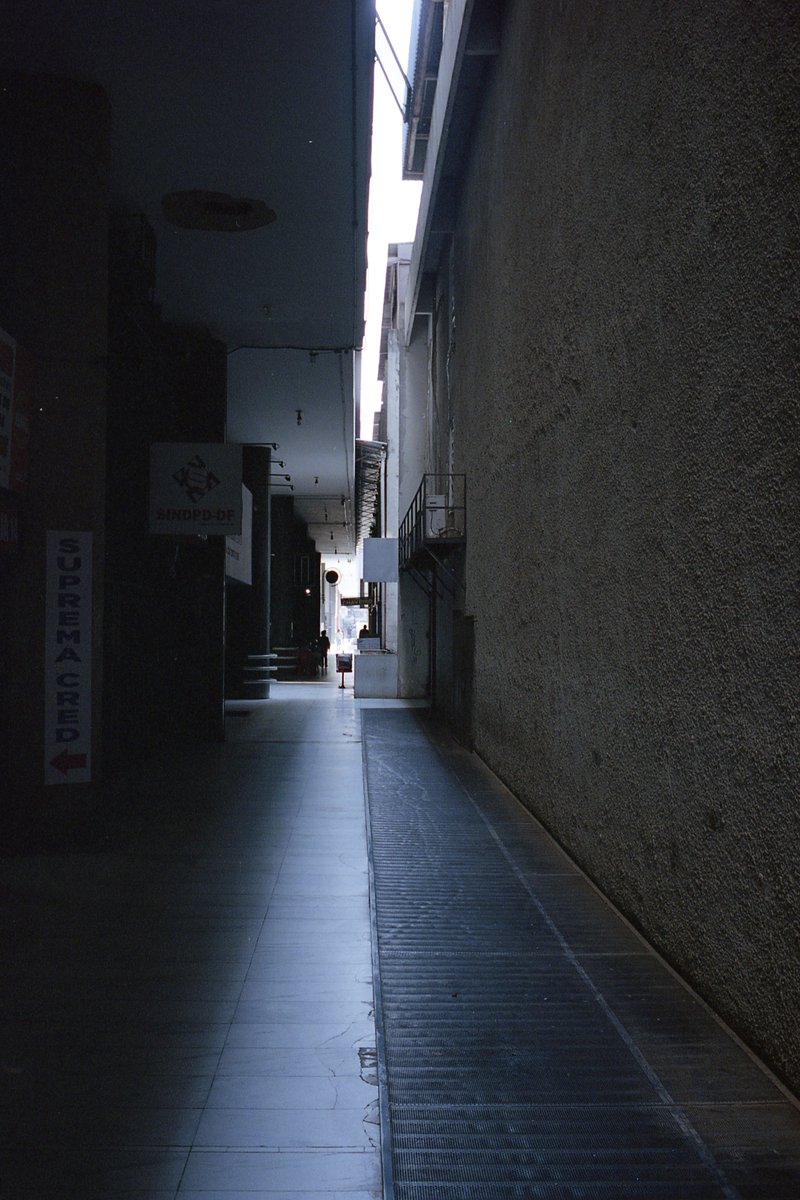 This is a black and white photo of an indoor corridor. The corridor appears to be made of cement or concrete and is lined on both sides by buildings.