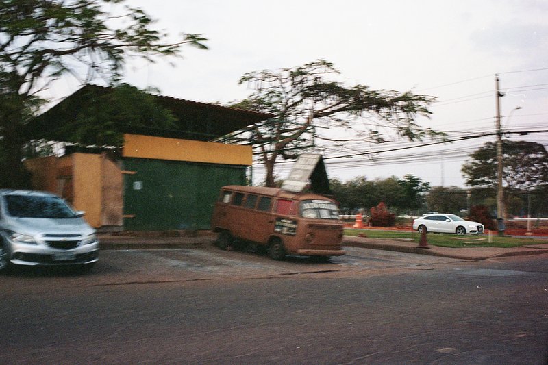 This is a photo of a small building with a thatched roof. In the foreground, there's an orange car parked near some trees and buildings. The backgroun