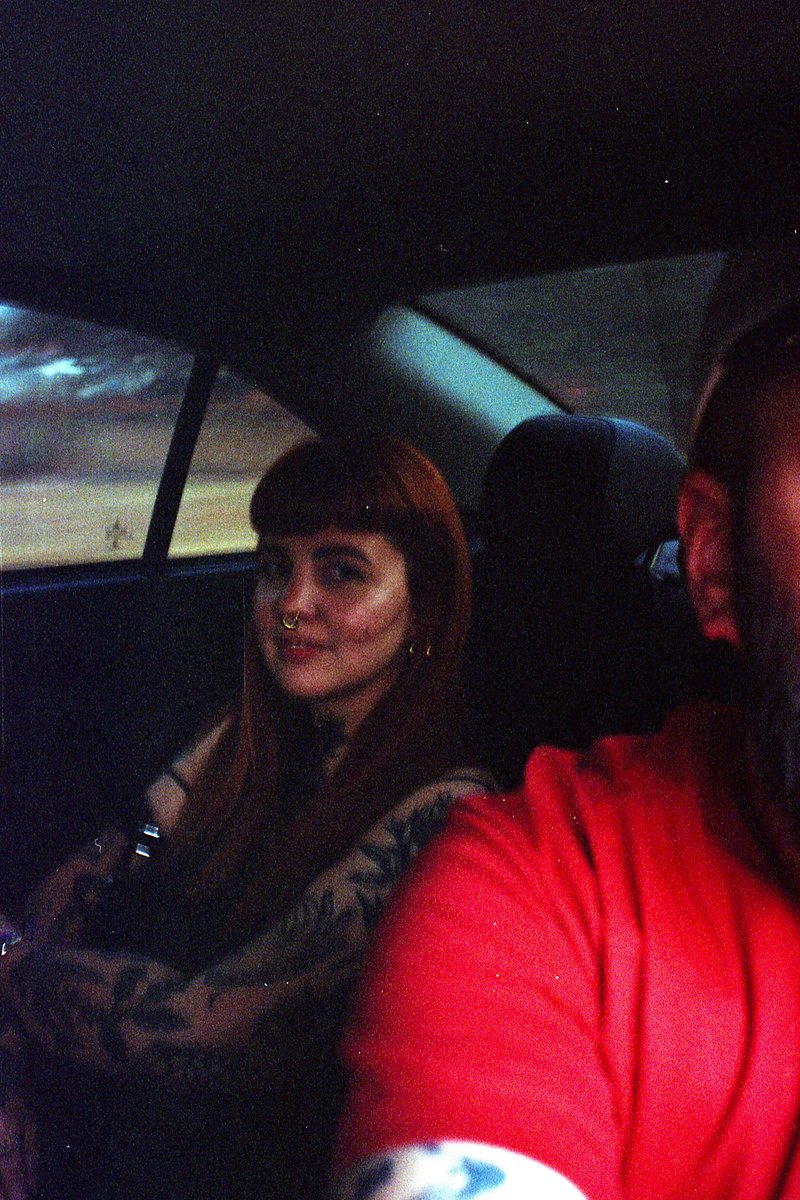 The image captures a unique moment of a person with tattoos and piercings sitting in the back seat of a car. They are dressed in a black outfit, possi