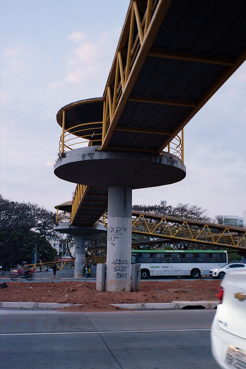This is a color photograph of a road bridge with yellow railings. The bridge has multiple arches and is set against a partly cloudy sky. It spans over