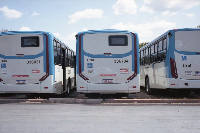 This is a photograph of three buses parked side by side in a parking lot. The buses are large, white vehicles with blue lettering and numbers on their