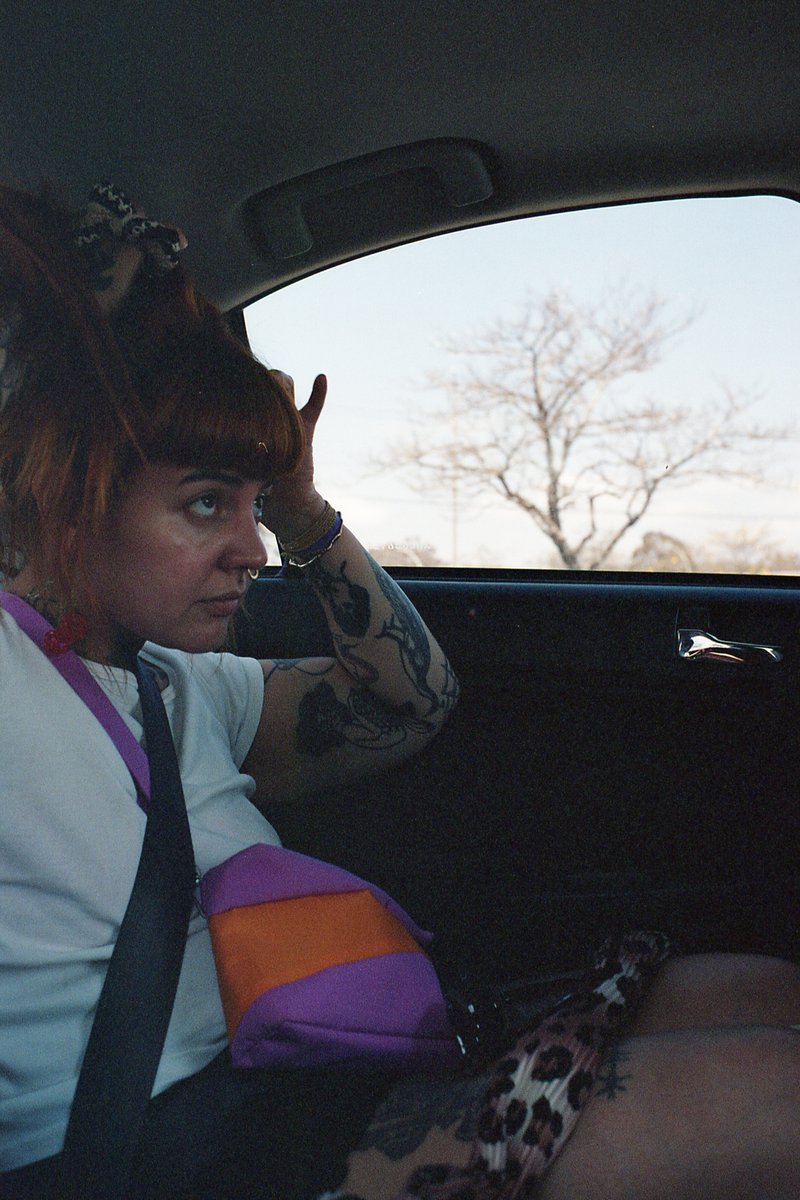 The image is a photo of a person in the foreground who appears to be seated inside a vehicle. The individual has red hair and is wearing a white t-shi
