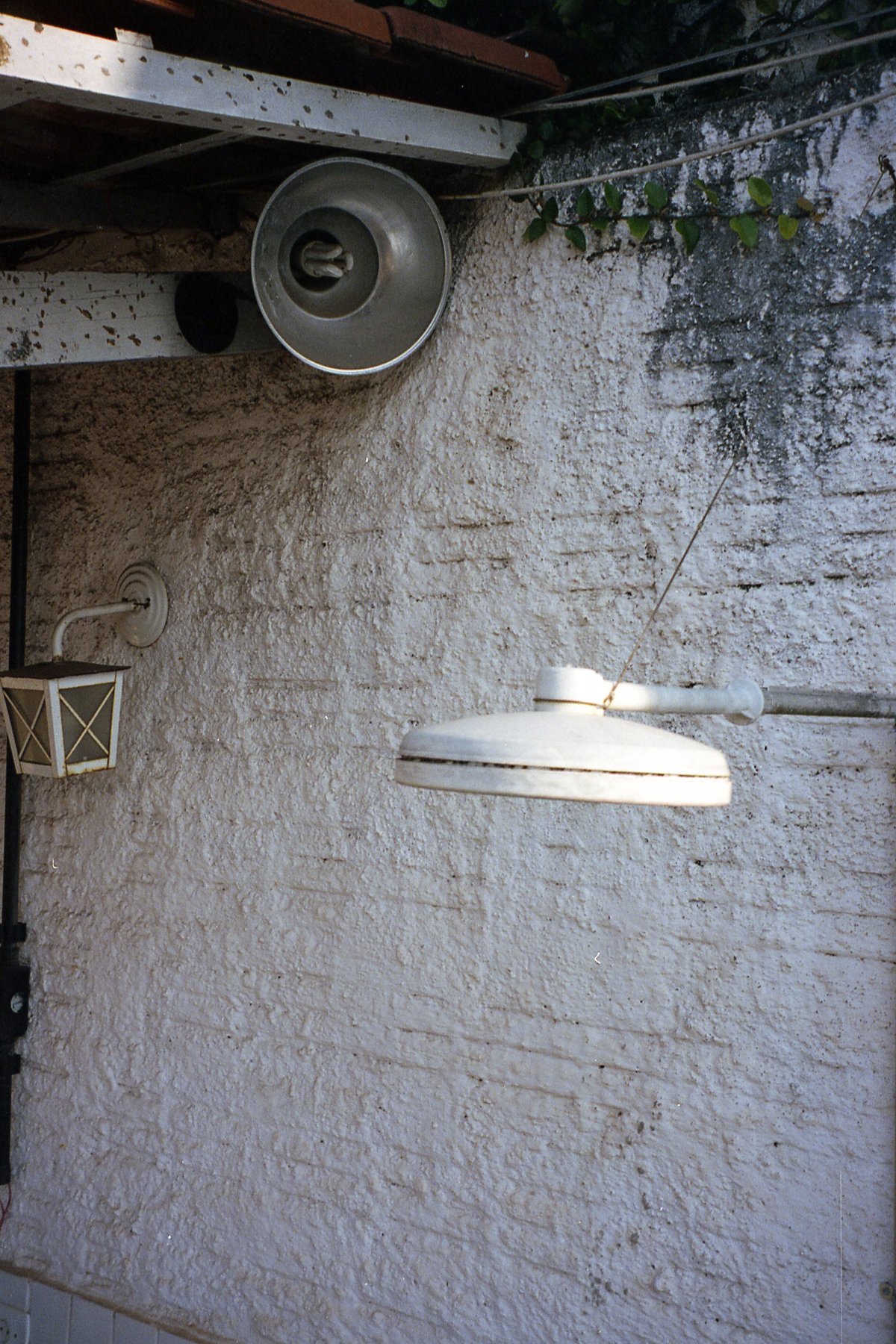 The image shows a white building wall with peeling paint. A rusted, unopened metal box is mounted on the wall. A hanging plant provides some greenery 