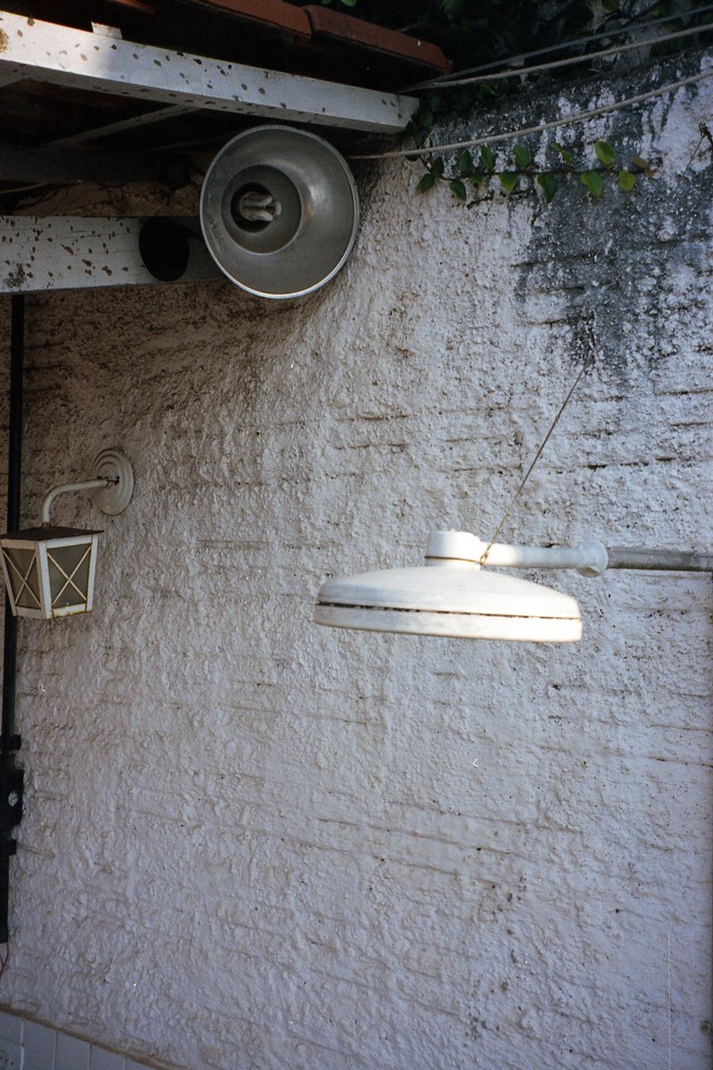 The image shows a white building wall with peeling paint. A rusted, unopened metal box is mounted on the wall. A hanging plant provides some greenery